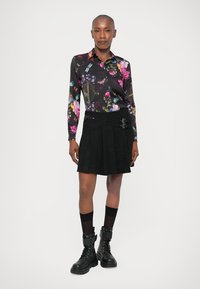 Egy színes virágmintás hosszú ujjú pólót, fekete pliszírozott szoknyát csattokkal, fekete zoknit és fekete fűzős katona nadrágcsizmát viselő nő áll fehér háttér előtt.