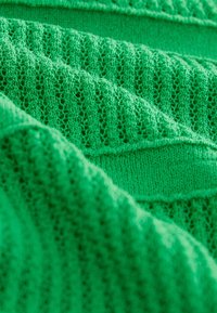 Grön stickad tyg med texturerade ränder och öppna vävmönster, som visar upp varierande stygnmönster och en slät, mjuk textur.