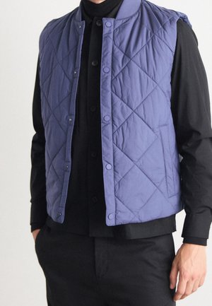 Homme portant une veste matelassée bleue par-dessus une chemise noire boutonnée et un pantalon noir, debout les mains dans les poches.