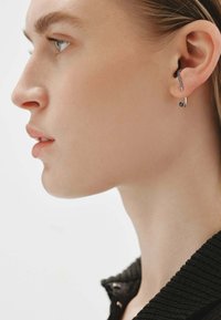 El perfil lateral de una mujer muestra un pendiente plateado con piedras azules a lo largo del borde de la oreja y una pequeña gema azul en el lóbulo, vistiendo una blusa negra texturizada.