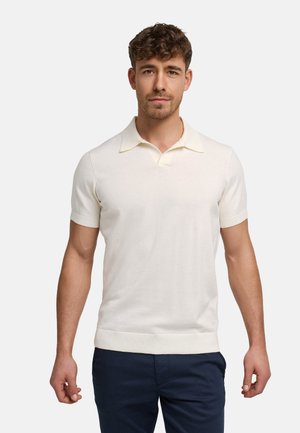 Jonge man met kort krullend haar, gekleed in een wit polo T-shirt met korte mouwen en een donkere broek, staand tegen een effen achtergrond.