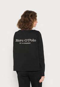 Černá mikina s volným střihem, dlouhými rukávy a žebrovanými manžetami. Na zádech je bílé text: "Marc O'Polo EST. IN STOCKHOLM."