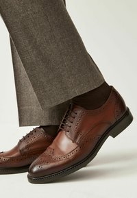 Pantofi brogue din piele maro cu perforații intricate, asortați cu șosete maro închis și pantaloni gri. Toc mic și finisaj neted.