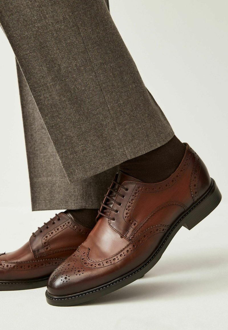 Pantofi brogue din piele maro cu perforații intricate, asortați cu șosete maro închis și pantaloni gri. Toc mic și finisaj neted.
