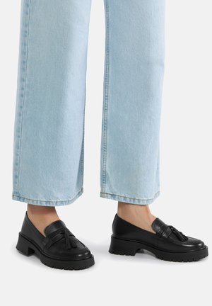 Pies con mocasines negros con borlas y tacones gruesos, combinados con vaqueros de pierna ancha y azul claro, recortados por encima de los tobillos, sobre un fondo blanco.