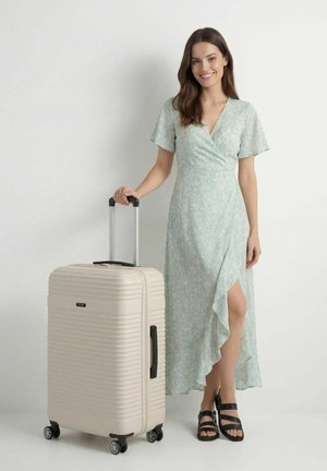 Uśmiechnięta kobieta w jasnej, zielonej sukience w kwiaty stoi obok beżowej twardej walizki z wysuniętą rączką na białym tle.