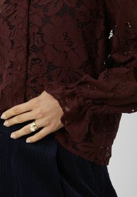 Top en dentelle bordeaux avec des motifs floraux, manches longues et poignets à volants. Associé à un tissu côtelé bleu marine en dessous, avec un accent en anneau doré.