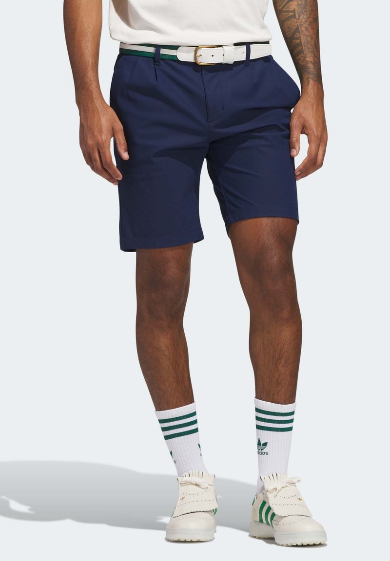 Marineblå shorts med flat front, med sidelommer. Kombinert med et hvitt belte med grønn aksent og et par hvite sokker med grønne striper.
