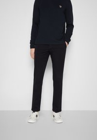 Svart långärmad skjorta med en liten logotyp, tillsammans med svarta raka byxor och vita sneakers med svarta detaljer och snörningen.