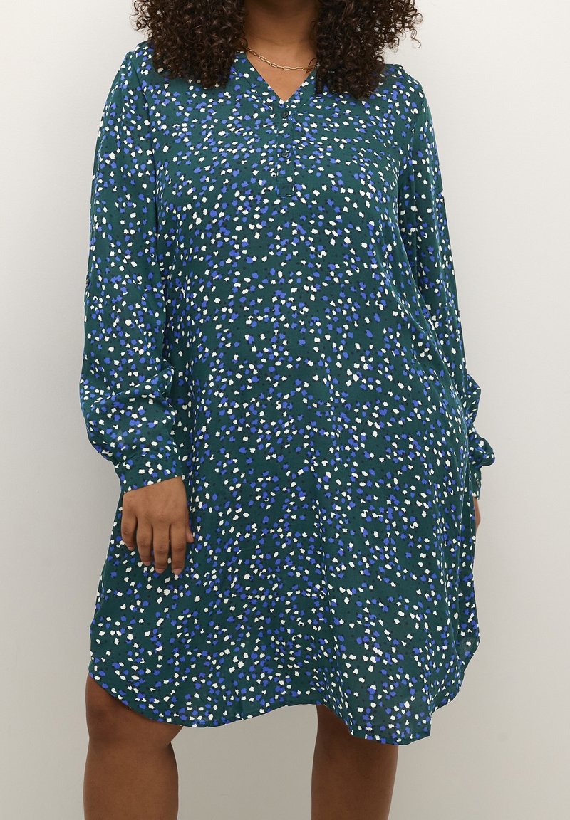 Femme portant une robe verte à manches longues, longueur genoux, avec un petit imprimé floral blanc et bleu, et un collier chaîne doré.