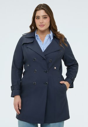 Mujer con abrigo trench azul marino de doble botonadura sobre una camisa azul con rayas, con una mano en el bolsillo del abrigo, mirando hacia adelante sobre un fondo liso.