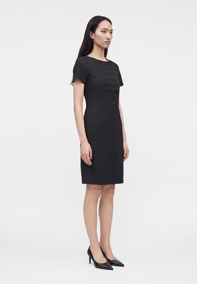 Femme portant une robe noire ajustée longueur genou, manches courtes et boutons décoratifs sur le devant, chaussée de talons noirs.