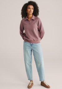 Svijetlo ljubičasta trenirka s ovratnikom i patentnim rubom, uparena s trapericama ravnih nogavica svijetlo plave boje i smeđim tenisicama s vezicama.