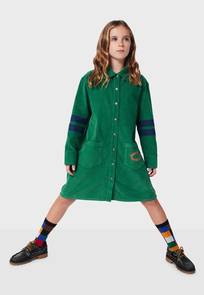 Grünes Cordkleid mit Knopfleiste, zwei vorderen Taschen, blauen Streifen an den Ärmeln und gesticktem Detail, kombiniert mit gemusterten Socken und Schuhen.