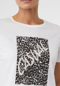 T-shirt blanc avec un motif abstrait en noir et blanc en forme de carré, comportant le mot "CASUAL" en beige. Manches courtes et col rond.
