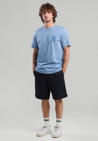 Vaaleansininen puuvillapaita, jossa on kukkakuva ja "CALIFORNIA" -teksti vasemmassa rinnassa, yhdistettynä mustiin shortseihin ja valkoisiin tennareihin.