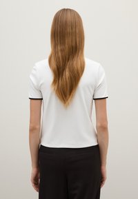 Top bianco a maniche corte con bordi neri, texture liscia, vestibilità slim e lunghezza leggermente accorciata. La parte posteriore è semplice, senza elementi di design visibili.