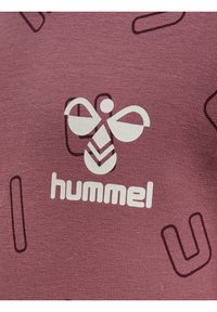 Maroon t-shirt med en mjuk textur, med en vit logotyp och ett abstrakt mörkt mönster. Framträdande varumärke i mitten av designen.
