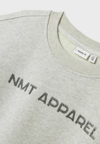 Felpa grigia con collo e polsini a coste, con "NMT APPAREL" stampato in lettere nere lucide sulla parte anteriore. Tessuto morbido.