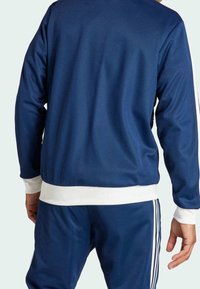 Blå sportjacka med vita detaljer, som har en mjuk yta, ribbade muddar och en figurnära passform. Designad för komfort och aktivt bruk.