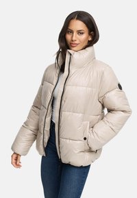 Giubbotto imbottito beige con colletto alto, chiusura a zip e tasche frontali. Design trapuntato con una texture liscia e lucida e dettagli in metallo nero.