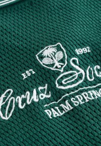 Grüner, strukturierter Stoff mit weißem, besticktem Text "Grand Soc" und "Palm Springs", einem Emblem mit einer Palme und Tennisschlägern sowie der Jahreszahl "1992".