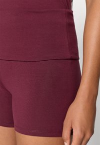 Κιμονό αθλητικού τύπου χρώματος μπορντό με εφαρμοστή σχεδίαση και ελαστικό ύφασμα, συνδυασμένο με  αντίστοιχες εφαρμοστές βερμούδες, αναδεικνύοντας μια ομαλή υφή και καθαρές ραφές.