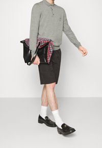Polo gris à manches longues, short noir, mocassins noirs et chaussettes blanches. Porte un sac noir avec une écharpe à carreaux rouge et bleue.
