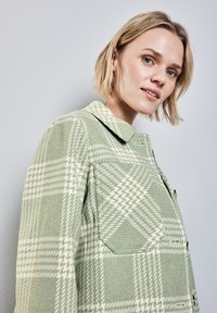 Zeleno-krémová kostkovaná bunda s límcem, která má knoflíkové zapínání a dvě náprsní kapsy. Vyrobena z texturované tkaniny s kostkovaným vzorem.