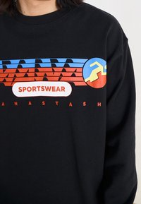 Zwarte sweatshirt met kleurrijke graphics in blauw, rood en geel, met het woord "SPORTSWEAR" duidelijk zichtbaar in wit.