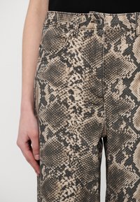 Gros plan sur le bas du torse d'une personne portant un pantalon taille haute avec un motif serpent beige et noir et une fermeture à bouton métallique.
