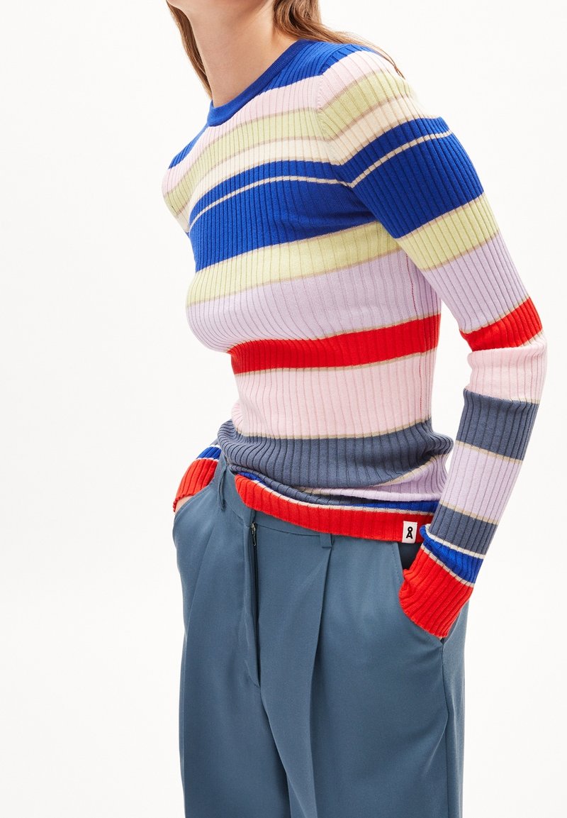 Personne portant un pull à rayures côtelées multicolores avec des rayures bleues, rouges, roses et grises et un pantalon bleu ample, les mains dans les poches.