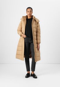 Lungo cappotto puffer beige con cappuccio e chiusura con zip, caratterizzato da sezioni trapuntate e una vestibilità rilassata. Abbinato a un top nero e jeans scuri.