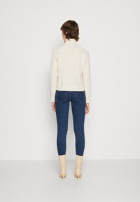 Pull en maille crème avec un col haut, associé à un jean skinny bleu. Bottines beige à bout pointu et surface texturée.