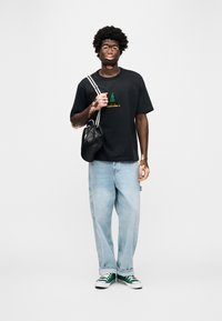 Μαύρη μπλούζα με σχέδιο δέντρου και πράσινες λεπτομέρειες, ανοιχτόχρωμο μπλε φαρδύ τζιν, πράσινα και λευκά αθλητικά παπούτσια, και μαύρη τσάντα με λευκές τιράντες.