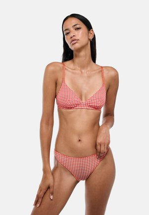 Femme aux longs cheveux foncés portant un bikini à carreaux rouge et blanc sur un fond uni gris clair.