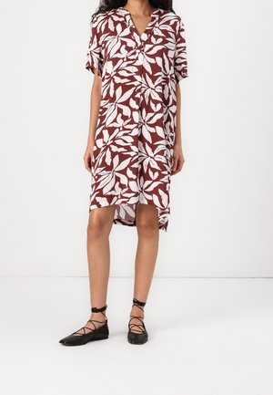 Femme portant une robe ample mi-longue aux genoux avec un imprimé feuilles bordeaux et blanc et des ballerines noires à lacets, debout devant un fond uni.