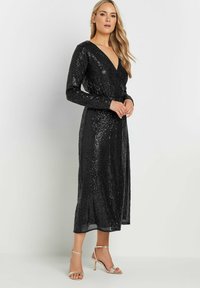 Svart paljettklänning med lång ärm, V-ringning och omslagdesign, med en flowy nederdel och åtsittande midja. I set med metalliska sandaler.