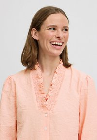Peach-colored blouse with a ruffled neckline and short puff sleeves. Textured fabric and front buttons add detail to the design.