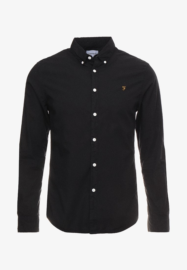 Chemise noire à manches longues boutonnée avec des boutons blancs, un col boutonné et un petit logo brodé en or sur la poitrine gauche.