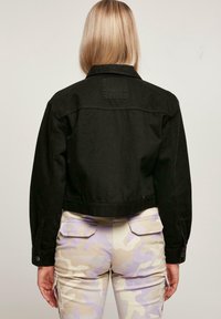 Svart kort denimjacka med spetsig krage och knäppta manschetter, matchad med lila byxor i kamouflage-mönster.