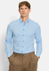 Light blue button-up shirt with dark polka dots, classic collar, long sleeves, and black buttons; paired with beige trousers.