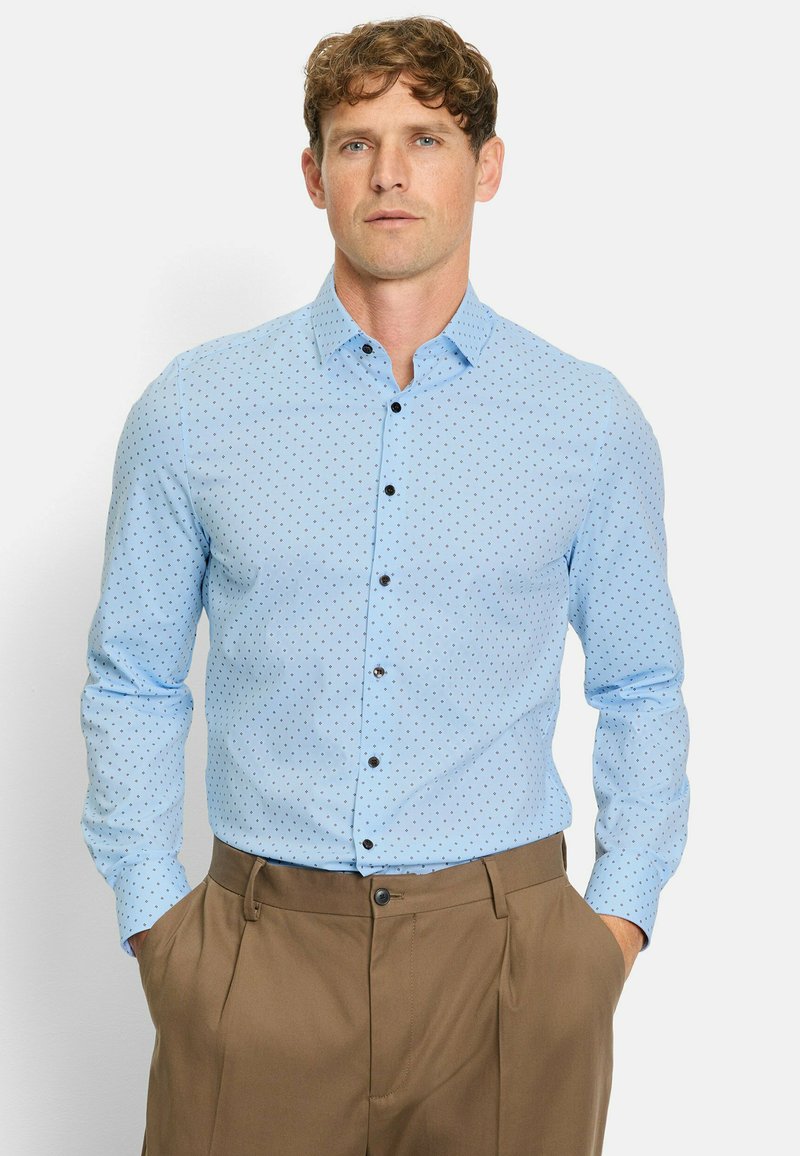 Light blue button-up shirt with dark polka dots, classic collar, long sleeves, and black buttons; paired with beige trousers.