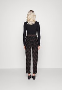 Kobieta zwrócona tyłem, ubrana w czarną bluzkę z długim rękawem, czarne spodnie z wysokim stanem ozdobione brązowym wzorem w zygzaki oraz czarne sandały na platformie.