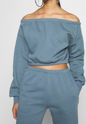 Femme portant un sweat-shirt bleu court à épaules dénudées et un pantalon de survêtement taille haute assorti, main dans la poche, boucles d'oreilles visibles.