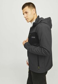 Zwarte softshell jack met een ritssluiting aan de voorkant, capuchon en gele accenten. Beschikt over een tweekleurig ontwerp en een logo op de borst.