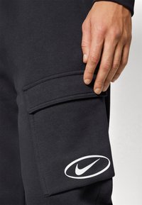 Svarta träningsbyxor med en sidoficka för cargo. Har en vit Nike-logotyp på det nedre benet. Materialet verkar mjukt och något strukturerat.