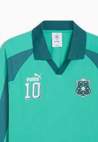 Maillot de sport Puma bleu sarcelle avec col foncé, numéro 10 sur le côté gauche de la poitrine, et un insigne complexe de style celtique sur le côté droit de la poitrine.