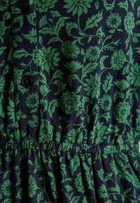 Tumedat sinine kangas, millel on roheline lillemuster, nupud ja klambriga talje detail. Pehme tekstuur lehe- ja lillemotiividega.