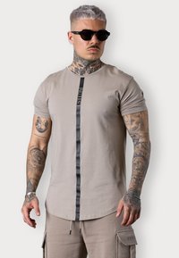 T-shirt beige à manches courtes avec une rayure verticale noire, arborant un texte de logo. Le modèle porte des lunettes de soleil, mettant en valeur des tatouages complexes sur les bras.
